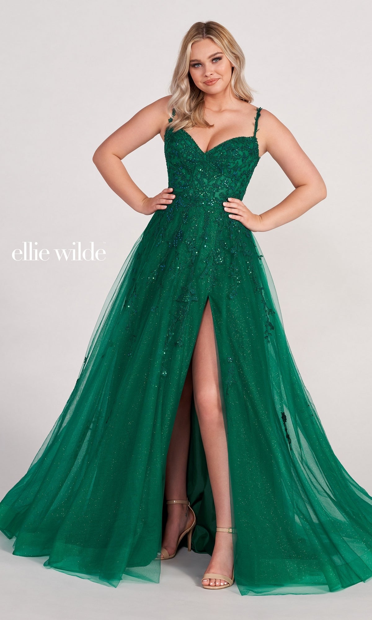 A woman poses against a plain background in the Ellie Wilde Long Glitter-Tulle A-Line Formal Dress featuring a sparkly emerald green fabric, sweetheart neckline, spaghetti straps, and a high slit.